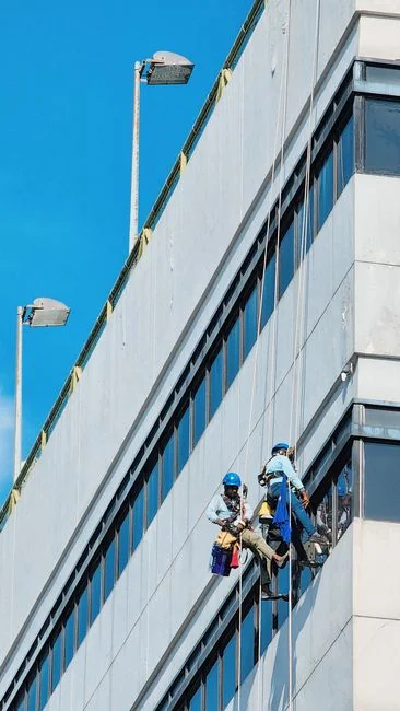 services-02 Black and white view from below of a steeplejack wearing a protective helmet and using professional gear to clean the glass wall of a modern tower